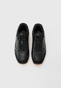 Une paire de baskets noires Armani Exchange avec des détails en cuir texturé et en daim, des lacets noirs et des semelles en caoutchouc brun sur un fond blanc.