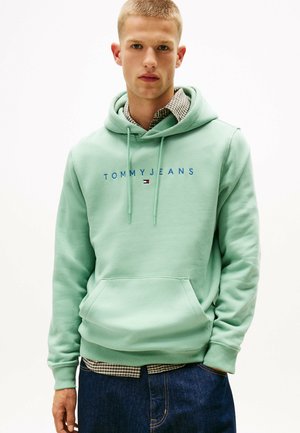 Mintgrüner Hoodie mit einer vorderen Taschenklappe, mit dem "TOMMY JEANS"-Logo in Blau. Wird über einem karierter Hemd getragen und mit blauen Jeans kombiniert.