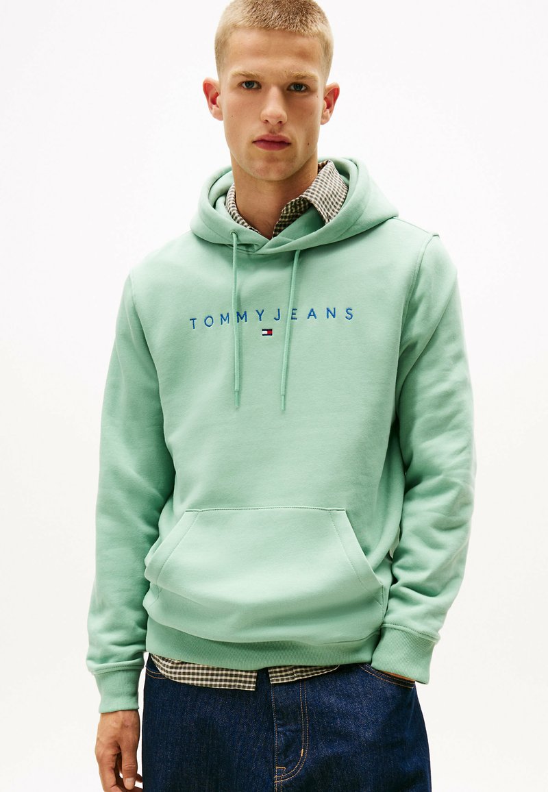 Mintgrøn hættetrøje med frontlomme, prydet med "TOMMY JEANS" logo i blå. Båret over en ternet skjorte og kombineret med blå jeans.