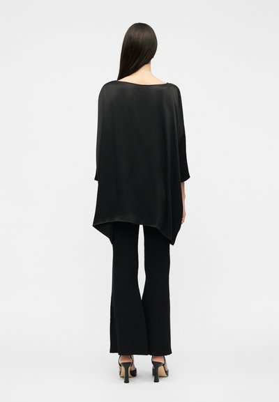 Žena s dugom tamnom kosom koja nosi široku crnu majicu, crne hlače na zvono i crne cipele s otvorenim prstima i visokim potpeticama, gledana s leđa.