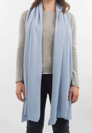 Femme portant une longue écharpe bleu clair sur un pull beige et un pantalon noir, debout devant un fond blanc uni.