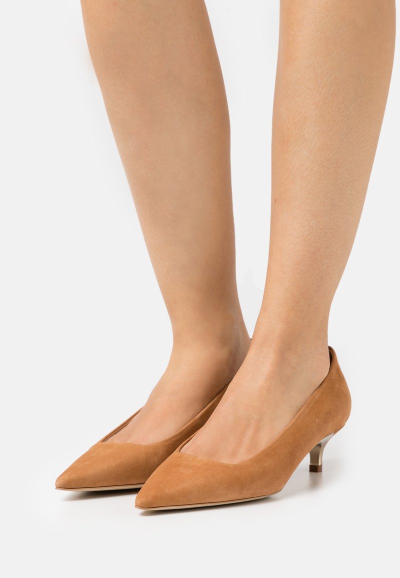 Jambes portant des escarpins pointus en daim beige avec de petits talons fins sur un fond blanc uni.