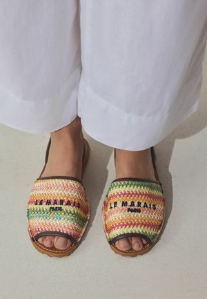 Fødder iført farverige vævede åbne tøfler med teksten "LE MARAIS PARIS", kombineret med løstsiddende hvide bukser, stående på lyst gulv.