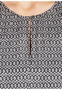Blusa negra con un patrón de diamantes en blanco. Abertura en forma de llave en el escote con un botón redondo. Textura de tela plisada.