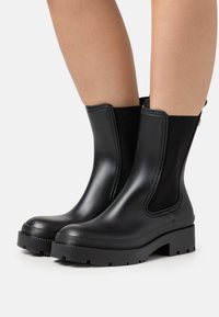 Stivali ankle neri in gomma con pannelli elastici laterali, suola spessa e strutturata, e finitura liscia, caratterizzati da un design minimalista e senza hardware visibile.
