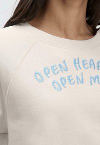 Sudadera beige claro con cuello redondo y mangas raglán. Presenta texto bordado en azul que dice "OPEN HEART OPEN MIND". Textura suave.