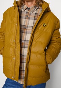 Padded hořčicově žlutá bunda s kapucí, vybavená zipovým zapínáním, bočními kapsami a malým vyšitým logem na hrudi.