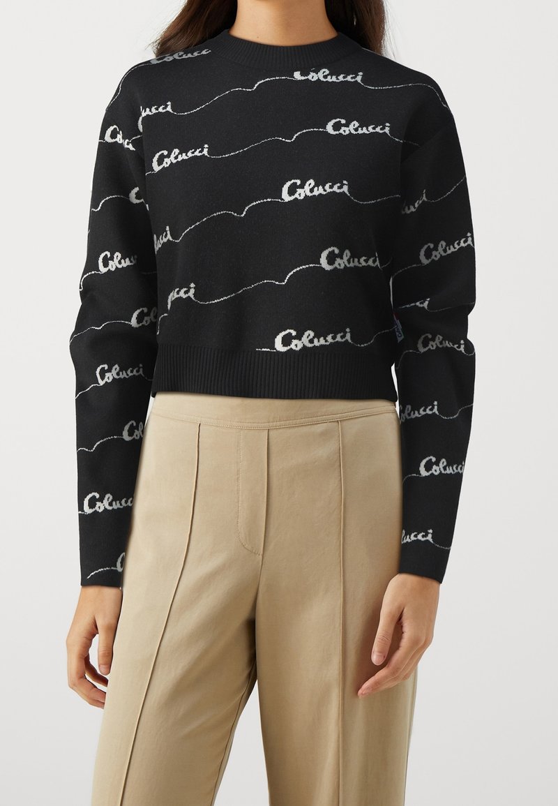Pull cropped noir à manches longues avec un motif script "Colucci" blanc, associé à un pantalon beige taille haute à plis.