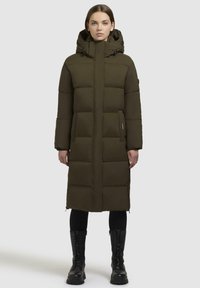 Long olive green puffer coat with a hood, featuring boxy quilted sections, a front zipper, and side pockets. Black knee-high boots worn underneath.