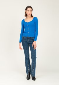 Geribbeld blauwe cardigan met een ronde halslijn en vijf goudkleurige knopen, gecombineerd met high-waisted blauwe jeans met knoopaccenten aan de zoom.