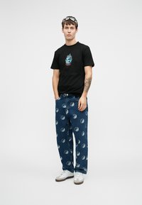 Μαύρη μπλούζα με γαλάζιο και κόκκινο γραφικό, συνδυασμένη με ανοιχτό γαλάζιο τζιν παντελόνι με μοτίβο γιν-γιαν σε όλη την επιφάνεια, λευκά αθλητικά παπούτσια.