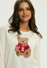 Kremowa koszula z długim rękawem z haftowanym misiem w czerwonym swetrze i kratce na szaliku oraz napisem "Ciesz się sezonem."