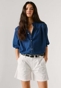 Donna con lunghi capelli scuri indossa una camicia di jeans blu abbottonata e pantaloncini bianchi con cintura nera, in piedi con le mani nelle tasche.