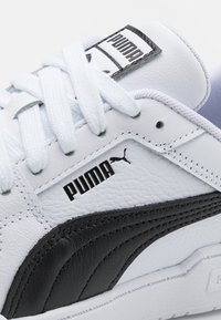 Baskets Puma en cuir synthétique blanc avec une bande d'accent noire. Elles présentent une finition texturée, des lacets plats blancs et un patch logo noir.