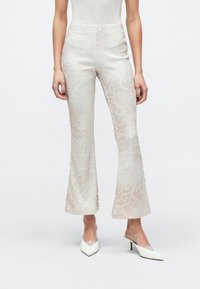 Υψηλοκάβαλα, καμπάνα παντελόνια σε ανοιχτό κρεμ με διακριτικό ροζ μοτίβο. Το ύφασμα φαίνεται λείο με απαλή υφή· συνδυασμένα με λευκά μυτερά παπούτσια.