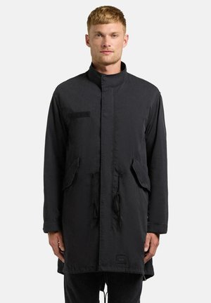 Mann mit kurzen blonden Haaren, der einen langen schwarzen Mantel mit hohem Kragen, Klappentaschen und Kordelzügen trägt, steht vor einem schlichten Hintergrund.