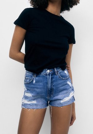 T-shirt en coton noir à manches courtes, associé à un short en denim bleu usé. Le short présente des ourlets effilochés et plusieurs déchirures.