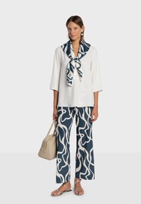 Haut blanc boutonné avec un foulard autour du cou, pantalon large à motifs bleu marine et blanc, sandales argentées à brides, et sac à main beige.