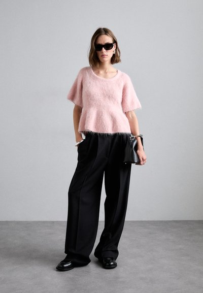 Růžový svetr z mohairu s krátkým rukávem, v kombinaci s černými širokými kalhotami. Modelka drží malou černou kabelku a nosí sluneční brýle.