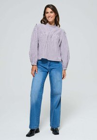 Femme souriante, portant un haut à col montant rayé avec des manches bouffantes, un jean droit bleu et des bottines à talons noires, se tenant sur un fond gris.