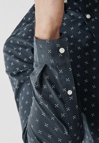 Donkerblauwe shirt met een knoopmanchet, voorzien van een patroon van kleine witte kruisen en stippen. Het materiaal lijkt textuur te hebben en is geweven.