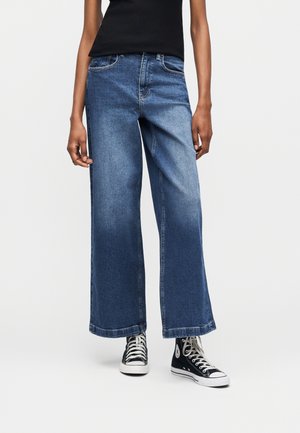 Άτομο που φοράει φαρδιά μπλε τζιν, μαύρο αμάνικο τοπ και μαύρα ψηλά αθλητικά παπούτσια, στέκεται μπροστά σε λευκό φόντο.