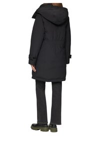 Zwarte gevoerde parka jas met een trekkoord in de taille, vaste capuchon en klittenbandsluiting bij de mouwen, gecombineerd met donkergrijze jeans en robuuste zwarte boots.