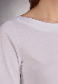 Maglia a maniche lunghe grigia chiara con scollatura ampia e tessuto liscio, leggermente testurizzato. I dettagli delle cuciture sono visibili nella zona delle spalle.
