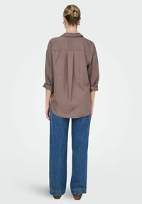 Brun skjorte med knapper og opfoldede ærmer, rygfold detalje, parret med løstsiddende blå jeans, vist fra ryggen.