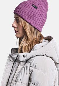 Beanie a costine viola con etichetta nera, abbinato a un giubbotto puffer grigio chiaro con colletto alto e chiusura con cordino.