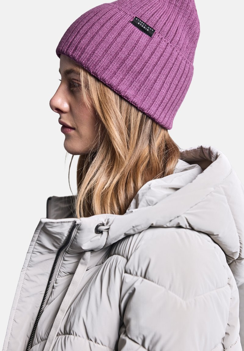 Beanie a costine viola con etichetta nera, abbinato a un giubbotto puffer grigio chiaro con colletto alto e chiusura con cordino.