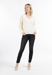 Crème V-hals trui met kleine gouden hartpatronen, gecombineerd met zwarte skinny jeans en beige hakken. Zachte gebreide textuur.