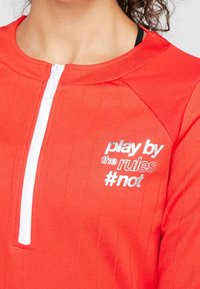 Haut de sport rouge à fermeture éclair avec une finition texturée, équipé de pièces métalliques blanches et d'un texte brodé : "joue selon les règles #not" en couleurs contrastées.