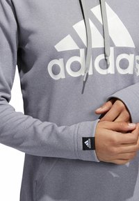 Sudadera gris con el logo blanco de Adidas, capucha con cordones y puños acanalados. Presenta una pequeña etiqueta negra de la marca en la manga. Textura de tela suave.