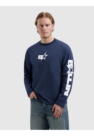 Jonge man met lichtbruin haar draagt een marineblauw shirt met lange mouwen met een witte ster en tekstprint op de borst en rechtermouw, gecombineerd met een blauwe spijkerbroek.