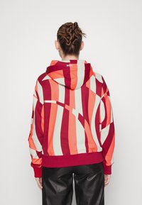 Sudadera a rayas rojas y naranjas con una gran capucha, diseñada en una tela suave. Presenta un logo en la parte posterior y puños y dobladillo acanalados.