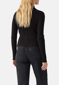 Schwarzes, geripptes Langarmoberteil mit einem Stehkragen, kombiniert mit dunklen Jeans, die ein kleines rotes Etikett an der Gesäßtasche haben. Glatte Textur, figurbetontes Design.