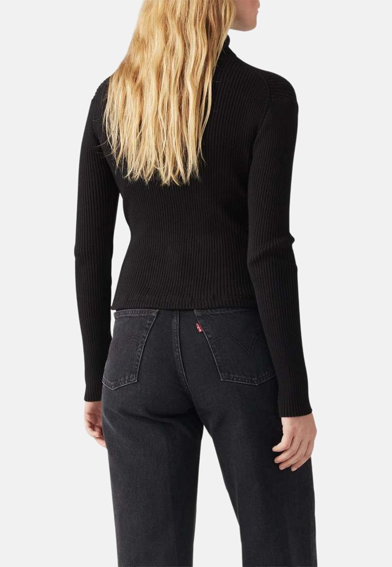Haut noir côtelé à manches longues avec un col montant, associé à un jean sombre avec une petite étiquette rouge sur la poche arrière. Texture lisse, design ajusté.
