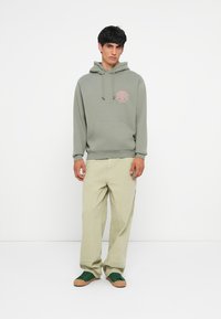 Jonge man staat in een saliegroen hoodie met logo, beige wijde broek en groene sneakers op een witte achtergrond.