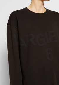 Dunkelbrauner Sweatshirt mit Rundhalsausschnitt, langen Ärmeln und großem schwarzem Textdruck mit der Aufschrift "MARGIELA 6" auf der Vorderseite. Weiches, strukturiertes Material.