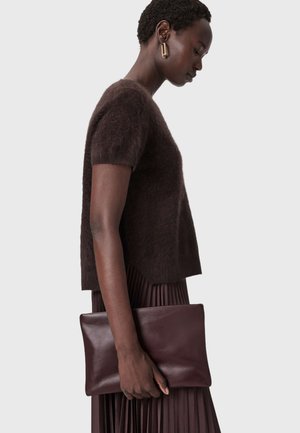 Brauner, flauschiger Strickpullover, kombiniert mit einem plissierten burgunderroten Rock. In einer Hand wird eine elegante burgunderrote Lederclutch gehalten, die ihre glatte Textur zeigt.