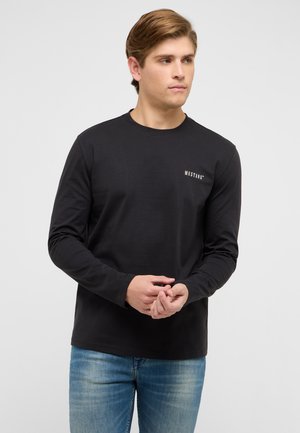 Junger Mann in schwarzem Langarmshirt mit "Mustang"-Logo auf der Brust, der die Hände richtet, blaue Jeans trägt und leicht nach rechts schaut, vor weißem Hintergrund.