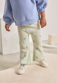 Pantalons à rayures vert clair avec des motifs floraux, ourlets effilochés et points noirs, associés à un sweat-shirt bleu et des baskets blanches.