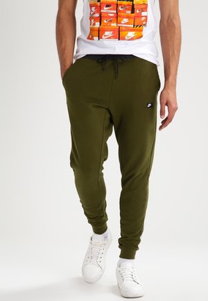 Person iført olivengrønne Nike joggers med sort snøre, hvide sneakers og en hvid t-shirt med stablet Nike skoboks grafik.