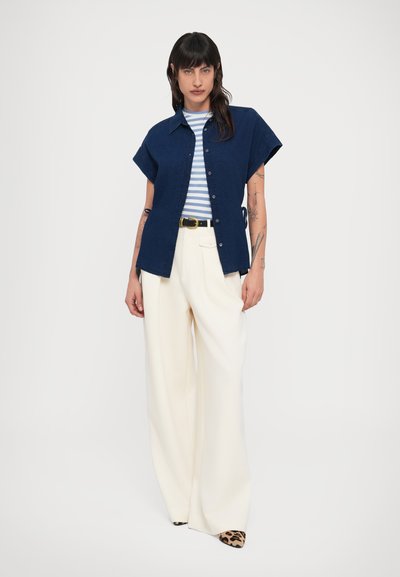 Πουκάμισο ναυτικού μπλε με κουμπιά και κοντά μανίκια, φορεμένο πάνω από λευκό και μπλε ριγέ μπλουζάκι. Συνοδεύεται από κρεμ παντελόνι με φαρδιά μπατζάκια και παπούτσια με animal print.