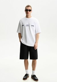 White oversized t-shirt with blue graphic text, black shorts, white socks, and black shoes with brown soles. Man stands against a light background.