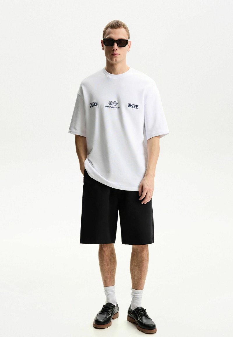 White oversized t-shirt with blue graphic text, black shorts, white socks, and black shoes with brown soles. Man stands against a light background.
