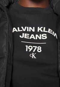 Sudadera negra con texto blanco que dice "CALVIN KLEIN JEANS 1978." Cuenta con un cuello acanalado y está combinada con una chaqueta acolchada negra.