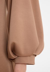 Manche en tissu beige avec un design à volants, se terminant par un poignet ajusté autour d'un bras à teint clair.