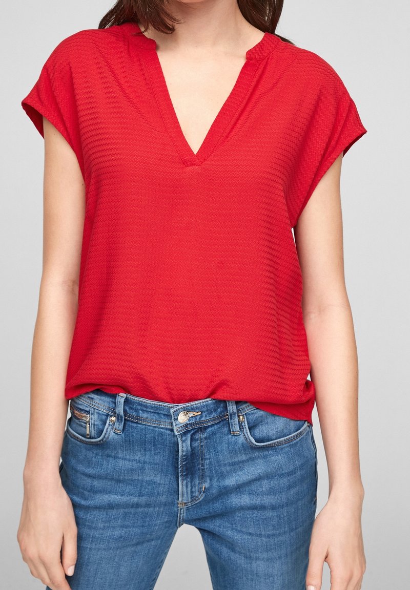 Blouse rouge texturée avec un col en V et des manches courtes. Associée à un jean en denim bleu avec poches avant et fermeture à bouton.
